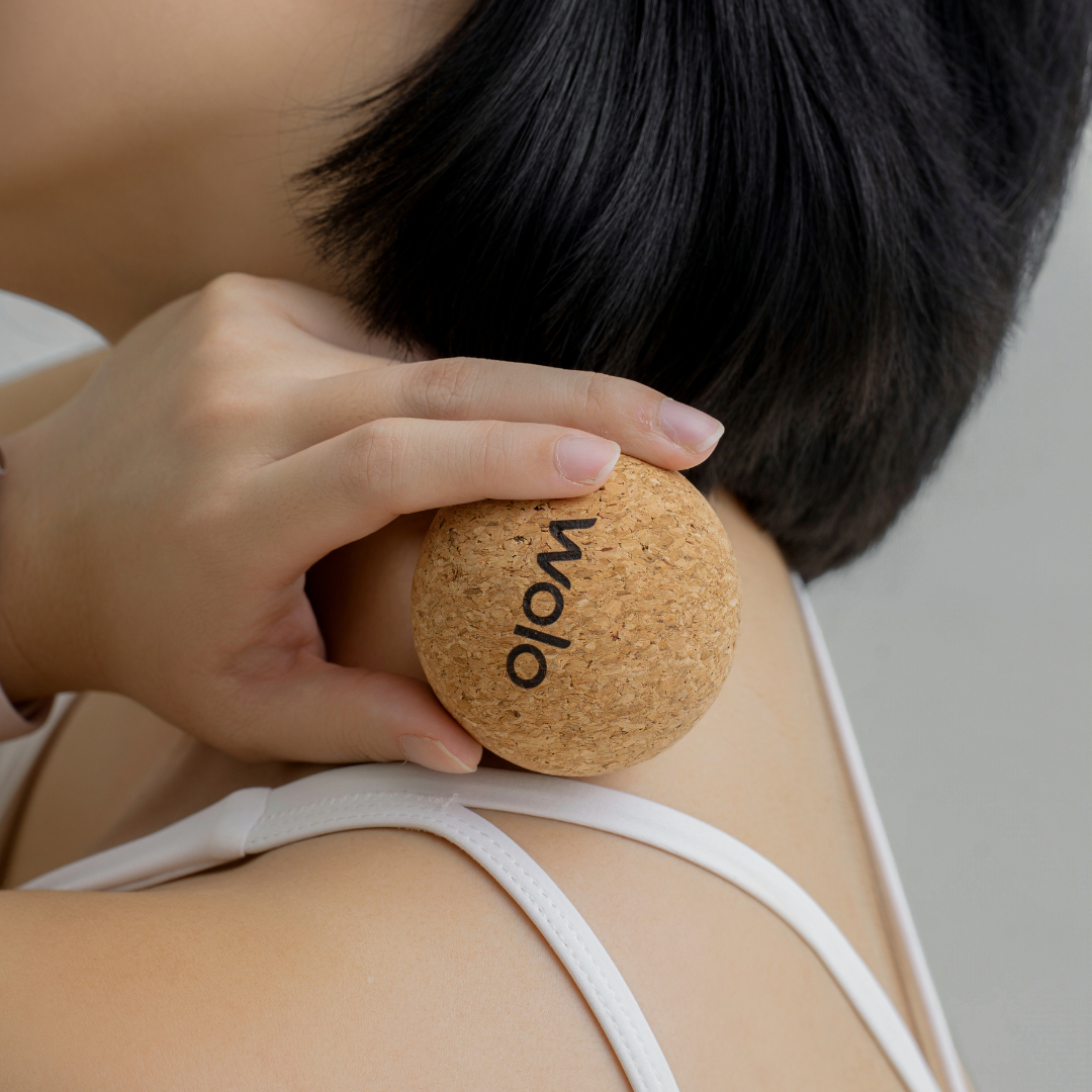 Person using a cork massage ball on her shoulder with 'Woo' branding.